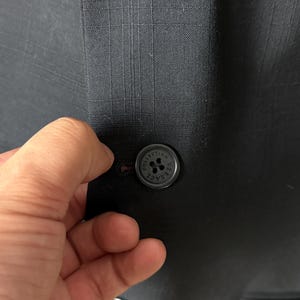 May include: Close-up of a dark gray blazer with a subtle plaid pattern. The blazer features a round, black button with the text "COLLECTION VERSACE" around the edge. The button is being fastened.