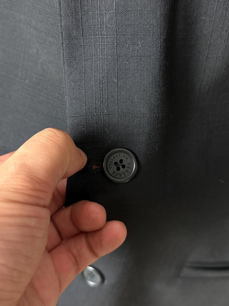May include: Close-up of a dark gray blazer with a subtle plaid pattern. The blazer features a round, black button with the text "COLLECTION VERSACE" around the edge. The button is being fastened.