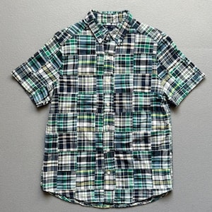 May include: A short-sleeved button-down shirt featuring a patchwork design composed of various green, navy blue, white, and yellow plaid fabric squares. The shirt has a standard collar, a front button placket, and a curved hemline at the bottom.