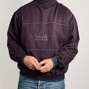 May include: A dark purple long-sleeve sweatshirt featuring a mock neck collar with a teal inner lining. The chest displays embroidered text reading The NATIONAL DIADORA EXPLORERS in red and teal. Teal stitching accents the chest and cuffs.