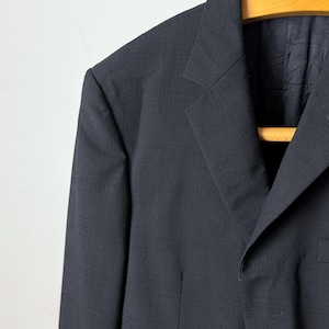 May include: Close-up of a black blazer hanging on a wooden hanger. The blazer has a subtle textured pattern and a notched lapel. The single-breasted jacket features a two-button closure and a chest pocket.