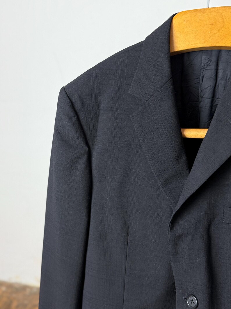 May include: Close-up of a black blazer hanging on a wooden hanger. The blazer has a subtle textured pattern and a notched lapel. The single-breasted jacket features a two-button closure and a chest pocket.