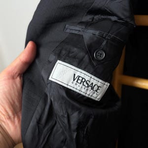 May include: Close-up of a black Versace Collection suit jacket. The interior lining is visible, with a label bearing the brand name. The jacket has a classic design with a button closure and a pocket detail. The fabric has a subtle textured pattern.