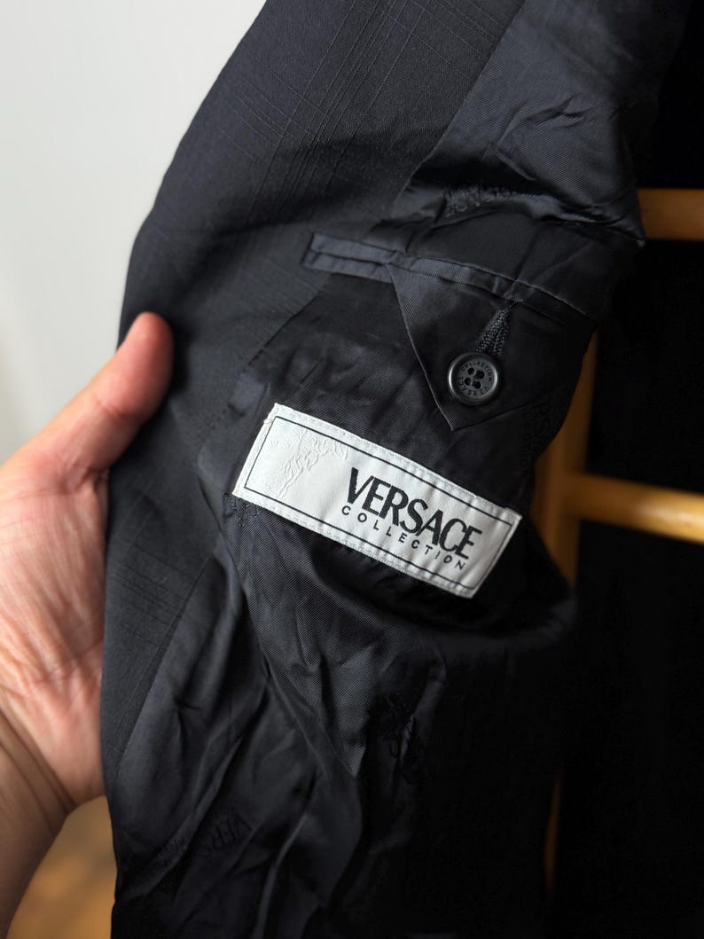 May include: Close-up of a black Versace Collection suit jacket. The interior lining is visible, with a label bearing the brand name. The jacket has a classic design with a button closure and a pocket detail. The fabric has a subtle textured pattern.