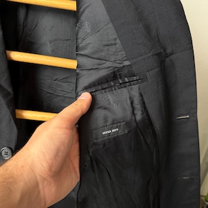 May include: Black blazer with a textured exterior and a black satin lining. The interior features a small pocket with the text "SUPER 100'S". The blazer is hanging on a wooden hanger.