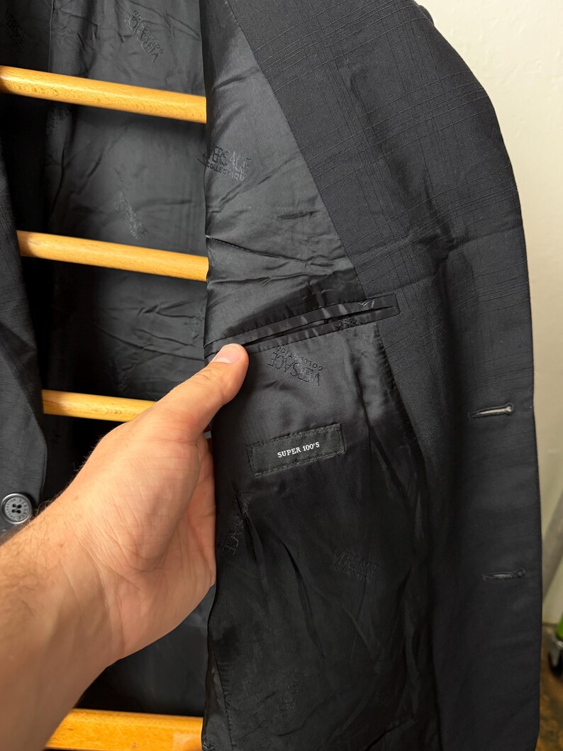 May include: Black blazer with a textured exterior and a black satin lining. The interior features a small pocket with the text "SUPER 100'S". The blazer is hanging on a wooden hanger.
