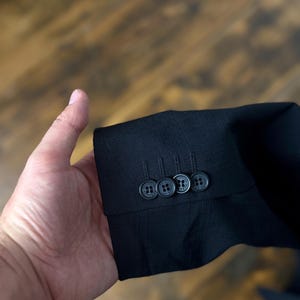 May include: Close-up of a black suit jacket sleeve with three matching buttons. The buttons are dark with a subtle pattern. The fabric has a textured appearance. The background is a blurred wooden surface.