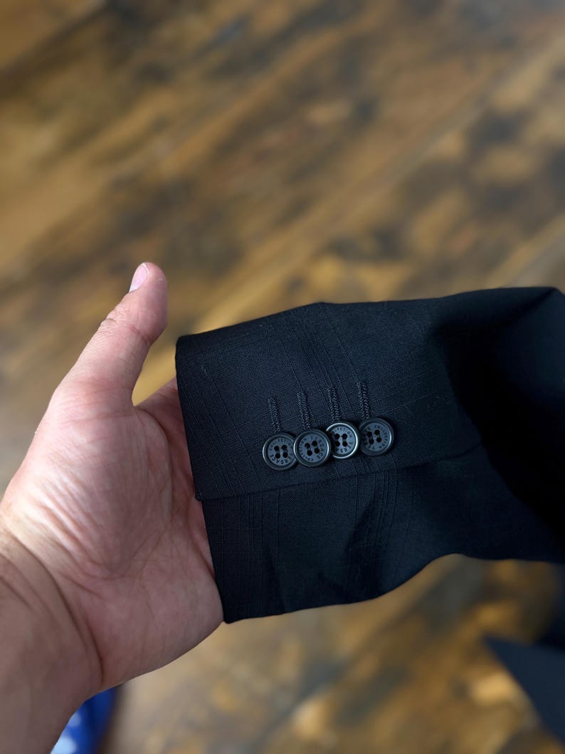 May include: Close-up of a black suit jacket sleeve with three matching buttons. The buttons are dark with a subtle pattern. The fabric has a textured appearance. The background is a blurred wooden surface.