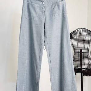 May include: Wide-leg trousers featuring a vertical pinstripe pattern in light blue and white. These pants include a front button closure, a waistband, and two rounded patch pockets on the front. The garment is displayed hanging on a wooden hanger.