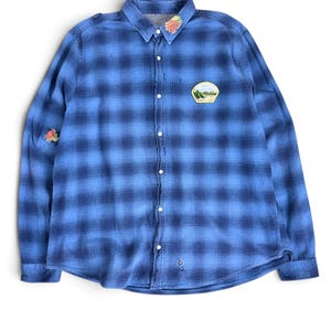 Pode incluir: Uma camisa de manga comprida com botões num padrão xadrez azul e azul escuro. A camisa apresenta um pequeno emblema de paisagem no peito esquerdo, um emblema de tecido colorido na gola e um pequeno emblema na manga direita.
