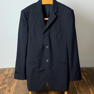 May include: A dark navy blazer with a three-button closure, hanging on a wooden hanger. The blazer has two front pockets and a notched lapel. The garment is displayed against a white wall and wooden floor.