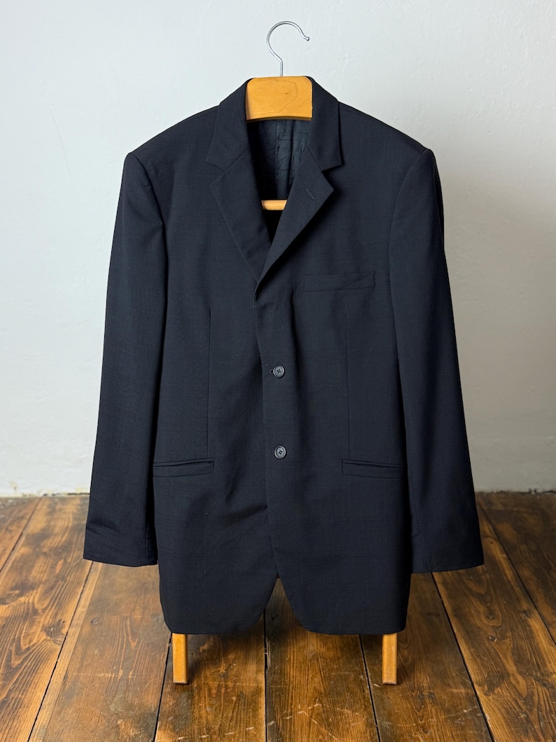 May include: A dark navy blazer with a three-button closure, hanging on a wooden hanger. The blazer has two front pockets and a notched lapel. The garment is displayed against a white wall and wooden floor.