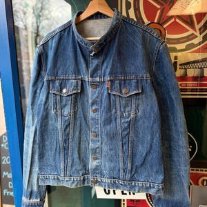 Puede incluir: Chaqueta vaquera azul vintage con cierre de botones, dos bolsillos en el pecho y cuello redondo. La chaqueta está colgada de una percha de madera. El logo de Levi's es visible en el bolsillo. Estilo clásico.