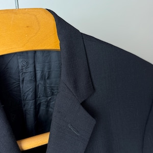 May include: Close-up of a black blazer with a subtle checkered pattern. The blazer is hanging on a wooden hanger. The interior lining is visible, with a brand label. The fabric has a textured appearance.