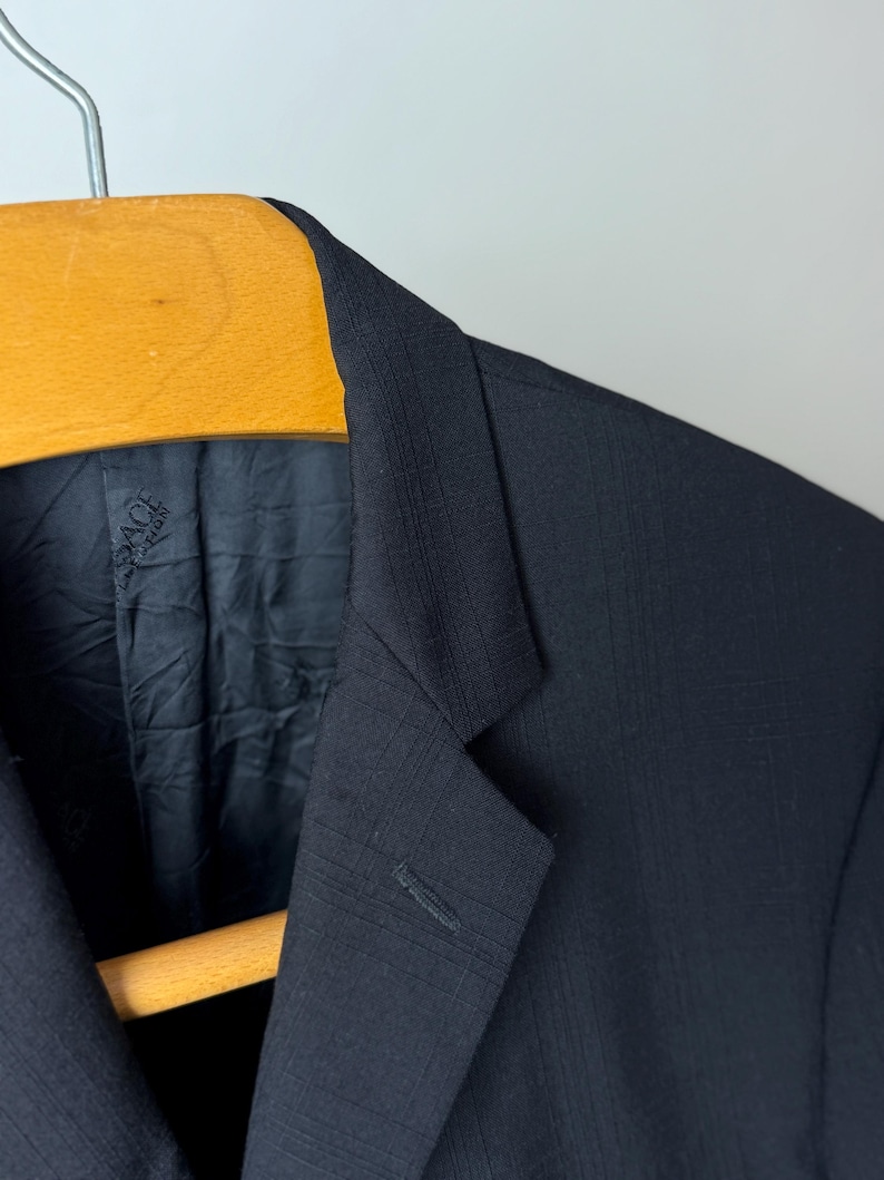 May include: Close-up of a black blazer with a subtle checkered pattern. The blazer is hanging on a wooden hanger. The interior lining is visible, with a brand label. The fabric has a textured appearance.