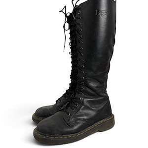 May include: A pair of black leather knee-high boots featuring a full-length lace-up front with black laces and metal eyelets. The boots have a thick, dark brown rubber sole with signature yellow stitching and a Dr. Martens logo embossed on the side.