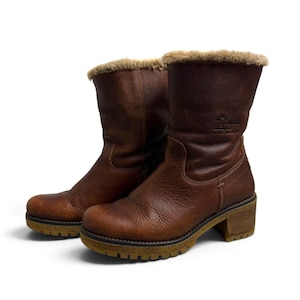 May include: A pair of mid-calf boots crafted from brown textured leather, featuring a soft beige faux fur lining at the collar. The boots are designed with a sturdy block heel and a thick, lugged rubber sole in a contrasting tan colour.