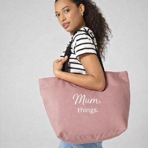 May include: A dusty rose tote bag with black straps and the words "Mum things." in white script. The bag is carried by a person wearing a black and white striped shirt.