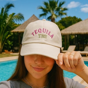 Könnte beinhalten: Beige Baseballkappe mit den Worten "TEQUILA TIME" in Rosa und Grün gestickt. Die Kappe wird von einer Person im Freien in der Nähe eines Pools getragen. Die Person trägt ein graues T-Shirt und goldene Halsketten.