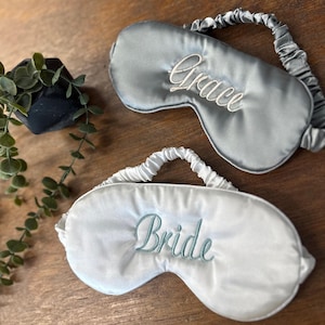 May include: Two satin sleep masks. One is grey with "Grace" embroidered in white. The other is white with "Bride" embroidered in light blue. Both have elastic headbands. The masks are on a wooden surface.
