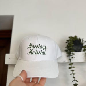 Puede incluir: Gorra de béisbol blanca con la frase bordada "Marriage Material" en verde. La gorra tiene una visera curva y está hecha de una tela lisa. El fondo es una pared blanca lisa.