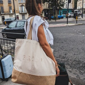 Op de afbeelding: Een vrouw draagt een grote beige tote bag met het woord "BRIDE" erop geborduurd. De tas heeft een bruine jute bodem en een lange band. Ze draagt ook een zwarte koffer.