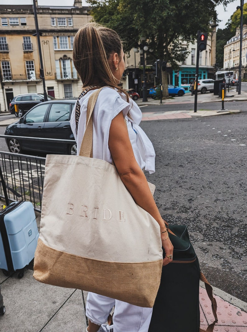 Op de afbeelding: Een vrouw draagt een grote beige tote bag met het woord "BRIDE" erop geborduurd. De tas heeft een bruine jute bodem en een lange band. Ze draagt ook een zwarte koffer.