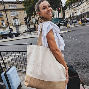 Op de afbeelding: Een vrouw draagt een tote bag met het woord "BRIDE" erop gedrukt. De tas is gemaakt van natuurlijke stof met een bruine jute bodem. Ze staat op een straat in de stad met een blauwe koffer en een zwarte koffer naast haar.