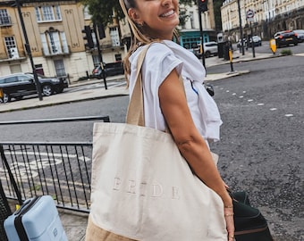 May include: A woman is carrying a tote bag with the word "BRIDE" printed on it. The bag is made of natural fabric with a brown burlap bottom. She is standing on a city street with a blue suitcase and a black suitcase beside her.