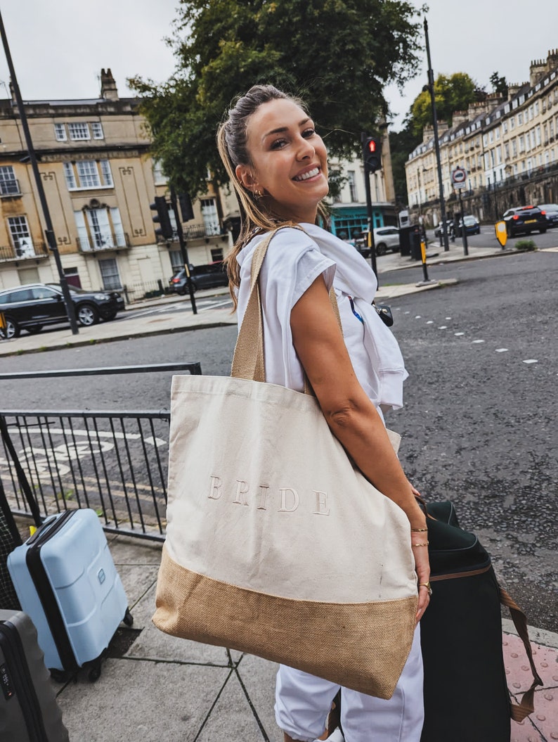 Op de afbeelding: Een vrouw draagt een tote bag met het woord "BRIDE" erop gedrukt. De tas is gemaakt van natuurlijke stof met een bruine jute bodem. Ze staat op een straat in de stad met een blauwe koffer en een zwarte koffer naast haar.