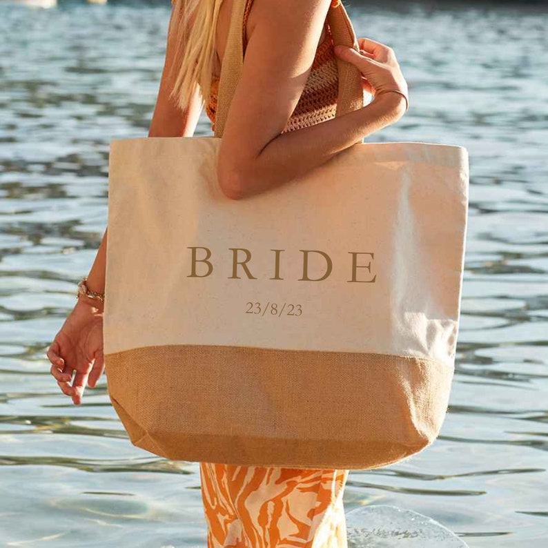 Op de afbeelding: Een witte canvas draagtas met een bruine jute bodem. De tas heeft het woord "BRIDE" in gouden letters gedrukt en de datum "23/8/23" eronder gedrukt.