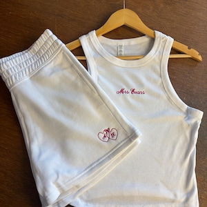 May include: White tank top and shorts set. The tank top features the embroidered text "Mrs Evans" in pink script. The shorts have an elasticated waistband and the embroidered initials "A & K" inside two hearts. The set is displayed on a wooden hanger.