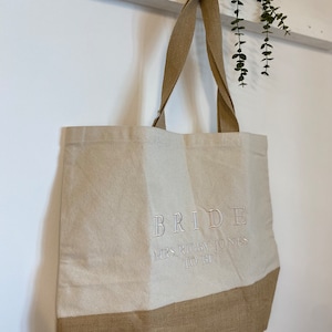 Op de afbeelding: Een beige tote bag met bruine jute bies. De tas heeft een wit geborduurd ontwerp met de tekst "BRIDE MRS RIGBY-JONES TO BE".