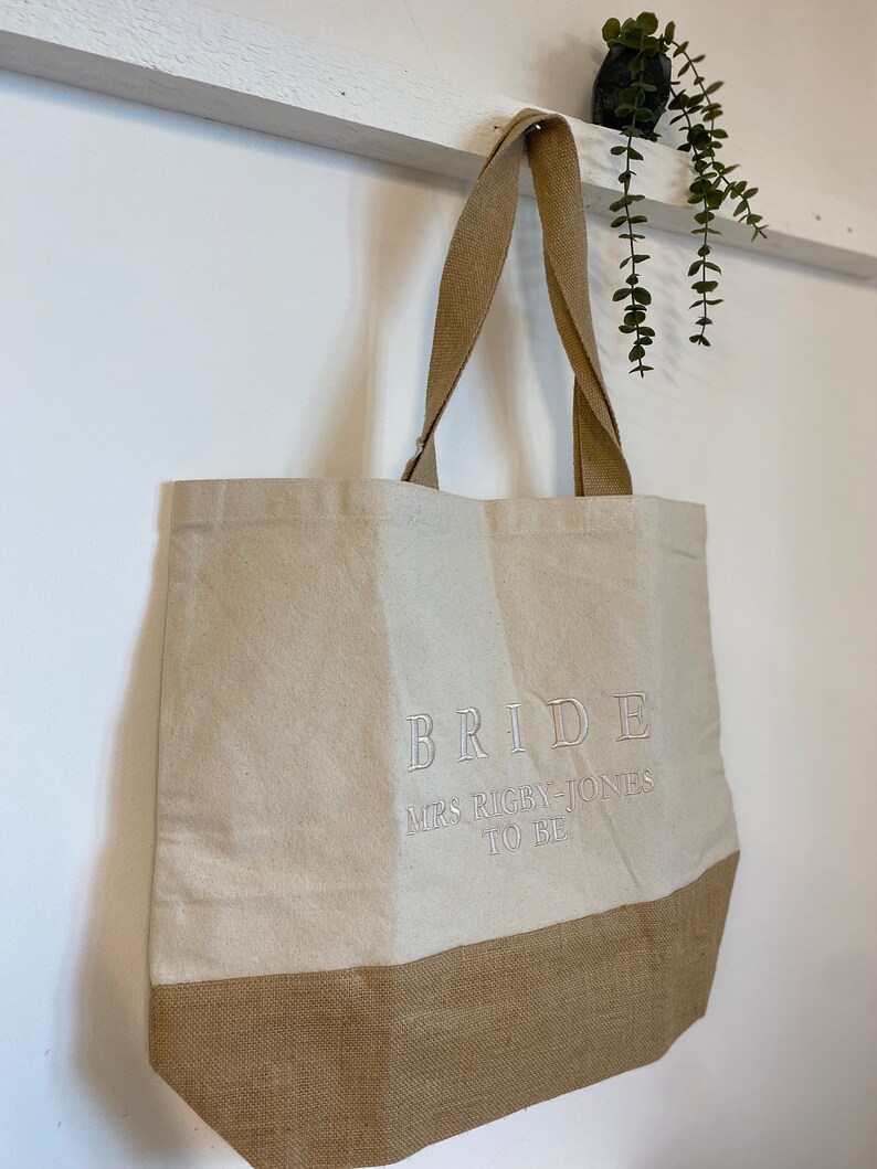 Op de afbeelding: Een beige tote bag met bruine jute bies. De tas heeft een wit geborduurd ontwerp met de tekst "BRIDE MRS RIGBY-JONES TO BE".