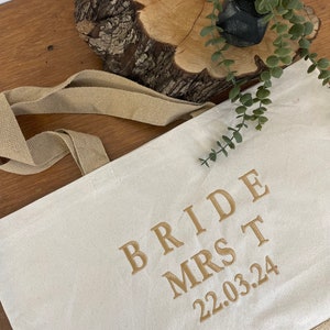 Op de afbeelding: Een witte canvas draagtas met bruine hengsels. De tas heeft gouden borduurwerk met de tekst "Bride Mrs T 22.03.24".