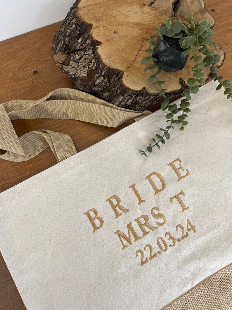 Op de afbeelding: Een witte canvas draagtas met bruine hengsels. De tas heeft gouden borduurwerk met de tekst "Bride Mrs T 22.03.24".