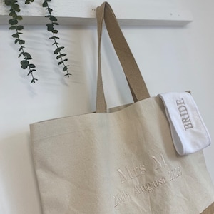 Op de afbeelding: Een beige canvas draagtas met bruine leren handvatten. De tas heeft een bruine jute bodem en is geborduurd met de tekst "Mrs M 26th August 2023". Een witte stoffen polsband met het woord "Bride" erop geborduurd is aan de tas bevestigd.