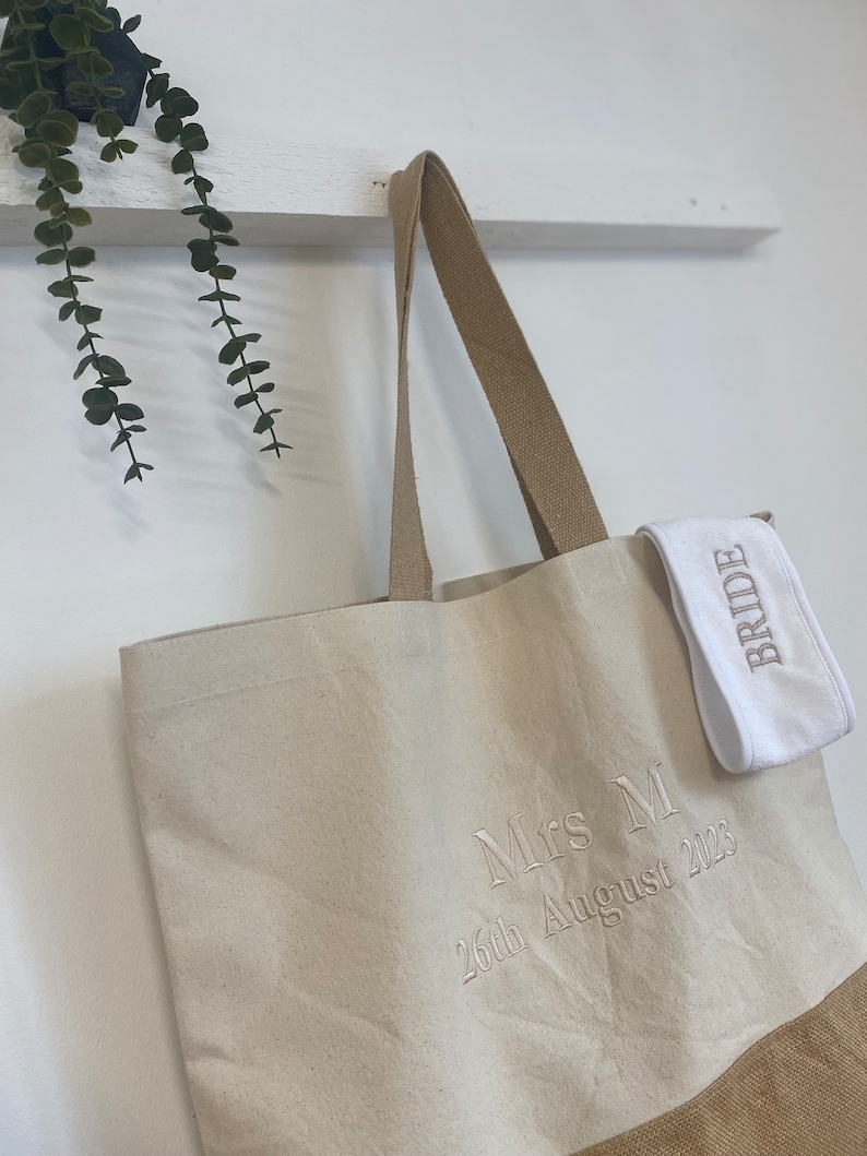 Op de afbeelding: Een beige canvas draagtas met bruine leren handvatten. De tas heeft een bruine jute bodem en is geborduurd met de tekst "Mrs M 26th August 2023". Een witte stoffen polsband met het woord "Bride" erop geborduurd is aan de tas bevestigd.