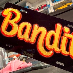 May include: A rectangular black license plate featuring the word Bandit in large, stylized yellow and orange gradient lettering. The text has a glossy finish with subtle highlights, and the plate includes four mounting slots at the corners.
