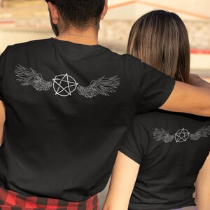 Könnte beinhalten: Zwei Personen tragen schwarze T-Shirts mit einem weißen Pentagramm- und Flügeldesign auf dem Rücken. Eine Person trägt ein rot-schwarz kariertes Hemd, die andere ein lila-grau kariertes Hemd.