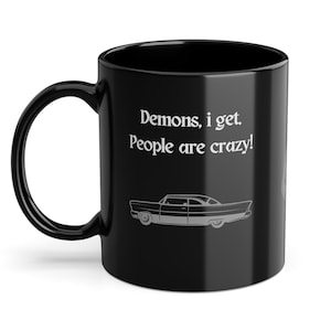 Könnte beinhalten: Schwarze Keramik-Tasse mit dem Text "Demons, I get. People are crazy!" und einer silbernen Auto-Grafik. Die Tasse hat einen gebogenen Henkel und eine glänzende Oberfläche. Ein lustiges Geschenk.