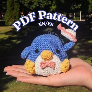 May include: Crocheted penguin amigurumi toy with a pink bow tie, yellow beak, and blue body. The penguin is wearing a pink and white crocheted hat. The text "PDF Pattern EN/ES @palupa_crochet" is visible in the image.