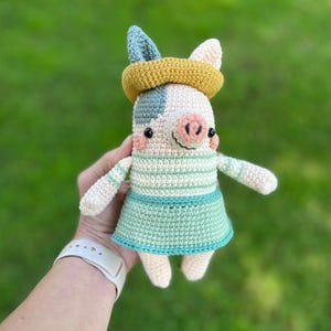 Könnte beinhalten: Handgemachte Häkelpuppe eines Schweins in Creme, Blau und Gelb. Das Schwein hat ein gestreiftes Oberteil, einen Rock und ein gelbes Stirnband. Die Puppe wird vor einem grünen Hintergrund gehalten.