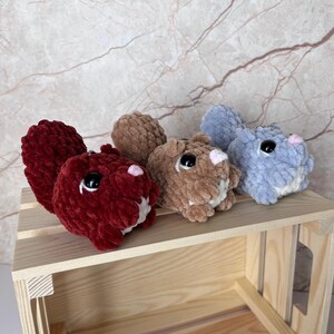 May include: Three plush squirrel keychains in shades of red, brown, and blue. Each squirrel has large black eyes, a pink nose, and a fluffy tail. They are displayed on a small wooden crate against a marble-patterned background.