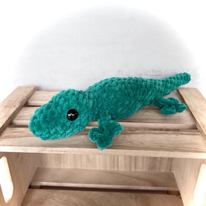 May include: A plush lizard toy crafted from teal-colored yarn with a soft, textured finish. The stuffed animal features a long tail, four small legs, and two smooth, round black plastic eyes positioned on the sides of its head.