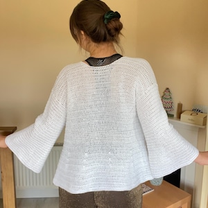 May include: White crochet cardigan with bell sleeves. The cardigan is loose fitting and has a slightly cropped length.