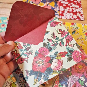Könnte beinhalten: Ein Papierumschlag mit einer dunkelroten Innenklappe und einer Außenseite mit einem lebendigen Blumenmuster aus rosa, roten und grünen botanischen Illustrationen auf cremefarbenem Grund, umgeben von weiteren Umschlägen mit bunten Blumenmustern.