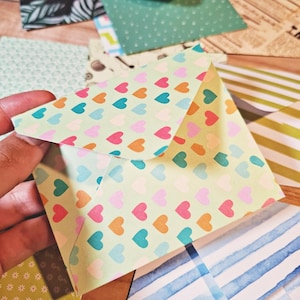 Könnte beinhalten: Eine Sammlung verschiedener Papierumschläge mit unterschiedlichen Mustern, darunter mehrfarbige Herzen, blau-weiße Aquarellstreifen, grün-weiße Blumenmuster, schwarz-weiße botanische Blätter und ein Druck im Vintage-Zeitungsstil.