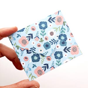 Könnte beinhalten: Ein kleiner, quadratischer Umschlag mit floralem Muster. Der Umschlag ist hellblau und mit einem sich wiederholenden Design aus rosa, weißen und blauen Blumen sowie grünen Blättern und Stielen versehen. Der Umschlag wird in einer Hand gehalten.