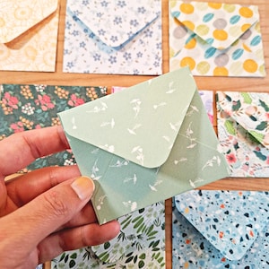 Könnte beinhalten: Ein kleiner, hellgrüner Papierumschlag mit einem zarten weißen Blumenmuster. Der Umschlag hat eine spitze Klappe und wird so gehalten, dass die Vorderseite sichtbar ist, während im Hintergrund weitere gemusterte Umschläge zu sehen sind.